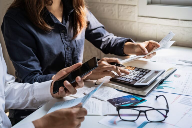 Mulher a fazer contas com calculadora, telemóvel e cartão bancário.