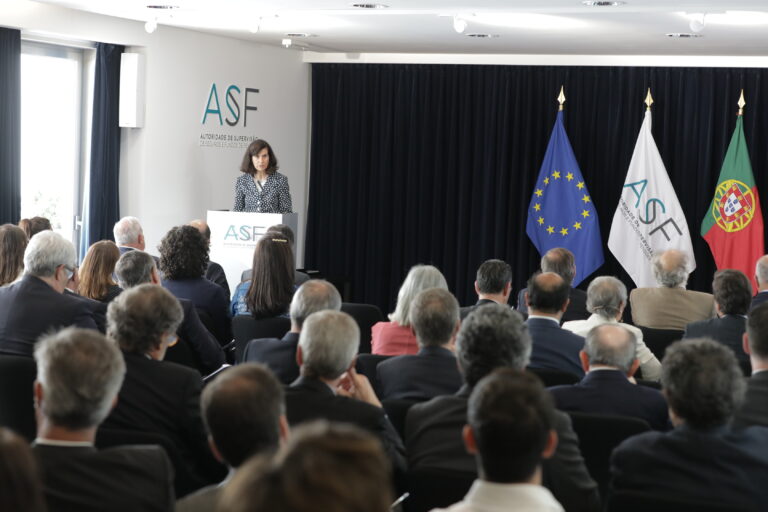A presidente da ASF fala para uma sala cheia e ao seu lado tem as bandeiras da União Europeia, da ASF e de Portugal.