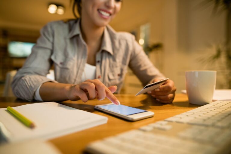 Mulher sentada ao computador segura um cartão bancário com uma mão e com a outra faz contas no telemóvel.