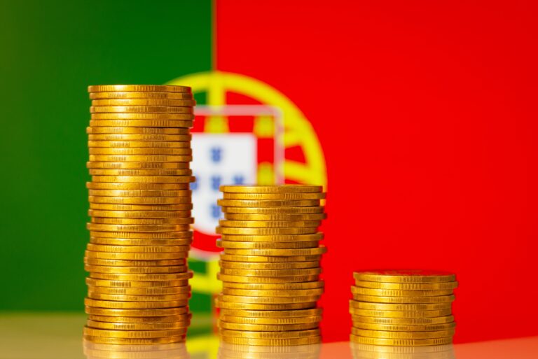 Três pequenos montes de moedas, de diferentes tamanhos, com a bandeira de Portugal ao fundo.