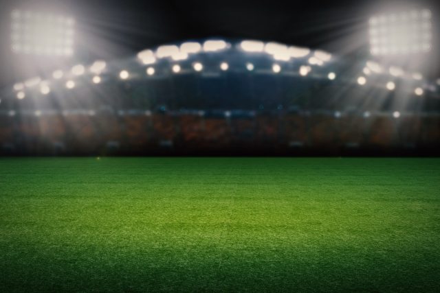 Relvado de um estádio de futebol à noite com bancadas em pano de fundo e as luzes acesas.