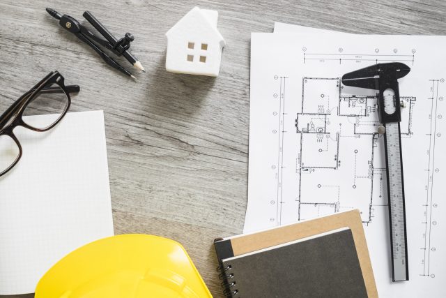 Projeto arquitetónico com cadernos, capacete de obras, casa em miniatura e outros instrumentos de desenho técnico.
