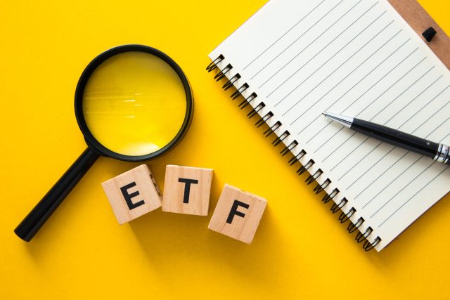Sobre um fundo amarelo está uma lupa, três quadrados de madeira que dizem ETF, um caderno em branco e uma caneta.